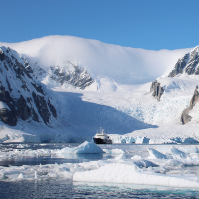 A small passenger vessel stands between icebergs with a spectacular view of the mountains of Antarctica