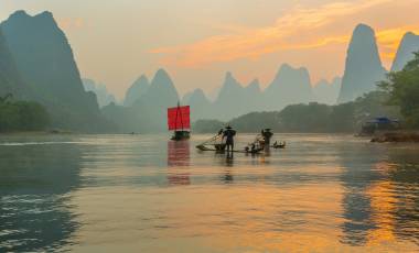 Fisherman stands on traditional bamboo boats at sunrise (boat with a red sail in the background)