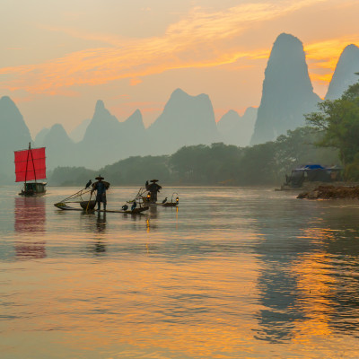 Fisherman stands on traditional bamboo boats at sunrise (boat with a red sail in the background)